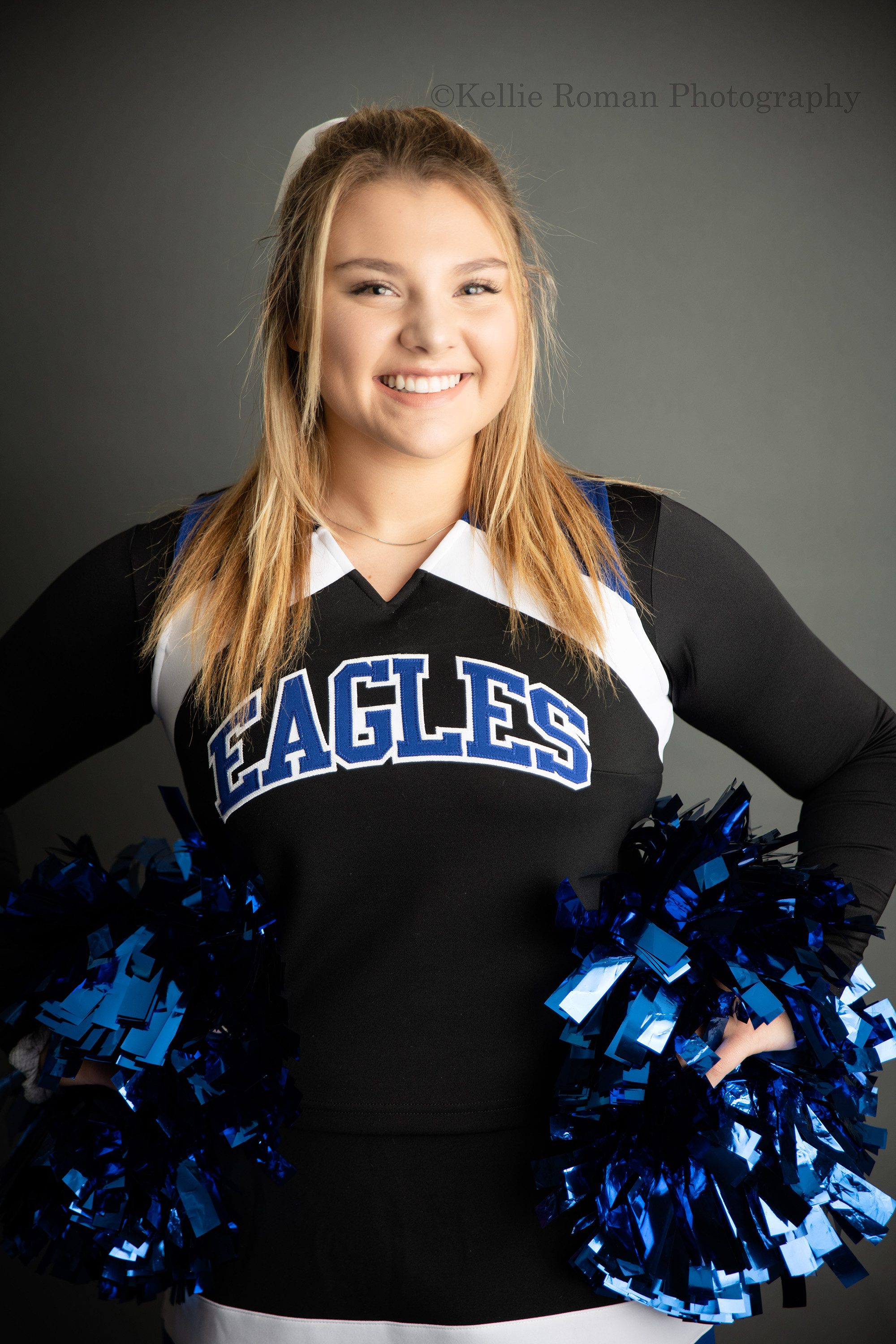 high school senior photographer a senior girl in a milwaukee photographers studio is wearing her cheerleading outfit. Her outfit is blue black and white. She's standing with her hands on her hips and her blue pom poms are in her hands.