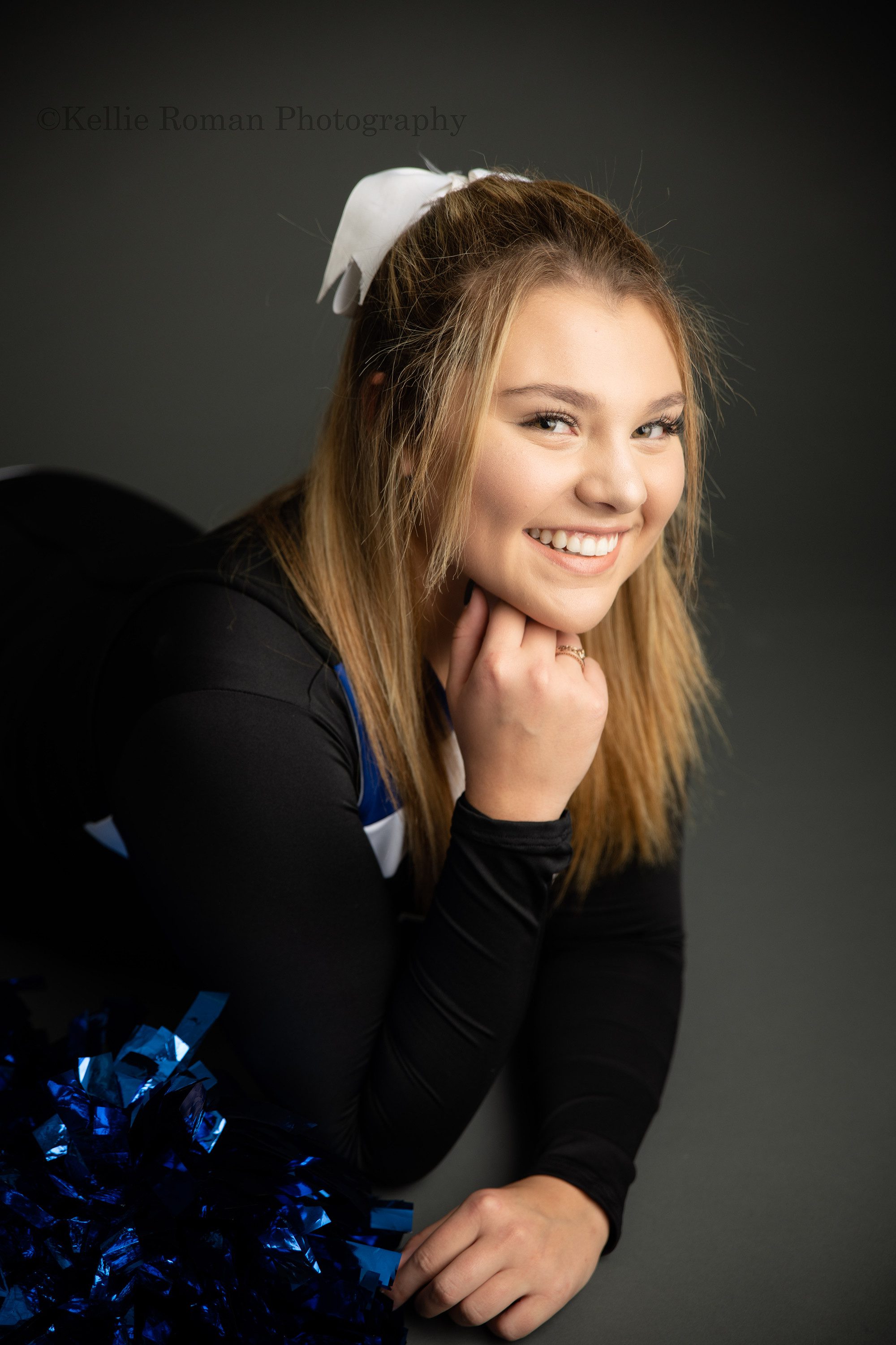 high school senior photographer a high school girl is in milwaukee photographers studio. The girl is laying on her stomach onto of a grey backdrop. She's wearing her blue and black cheer uniform and has her blue pom poms resting next to her. Her hand is under her chin.