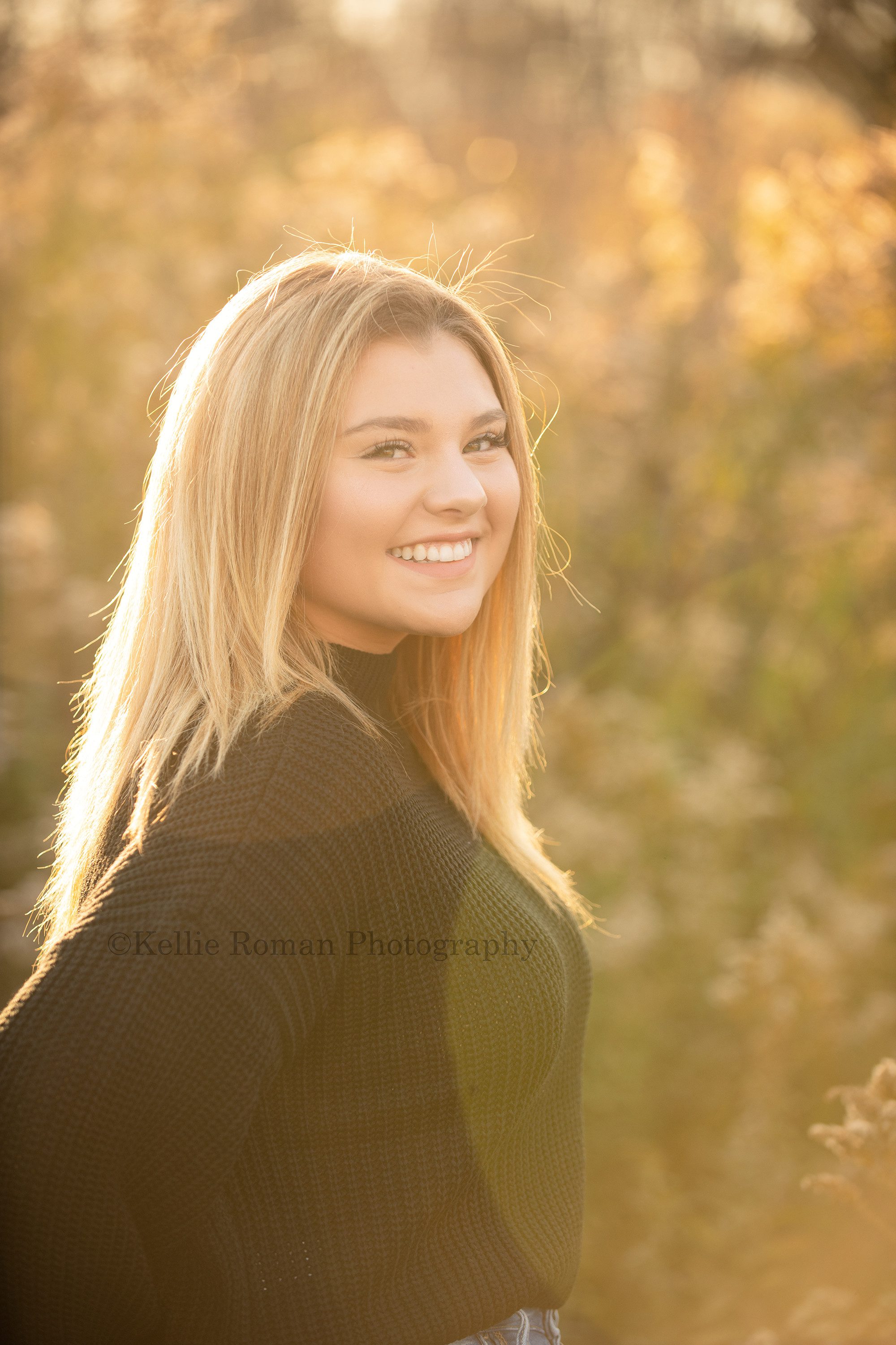high school senior photographer a blonde high school senior girl who's wearing a black sweater is standing in milwaukee county park. She has the sunset behind her and there is lots of golden sunlight through her blonde hair.