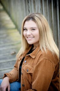high school senior photographer a high school senior girl is sitting on a wood bridge and leaning against the railing in a milwaukee county park she has on jeans and a brown jacket she has blonde hair and is looking at the camera