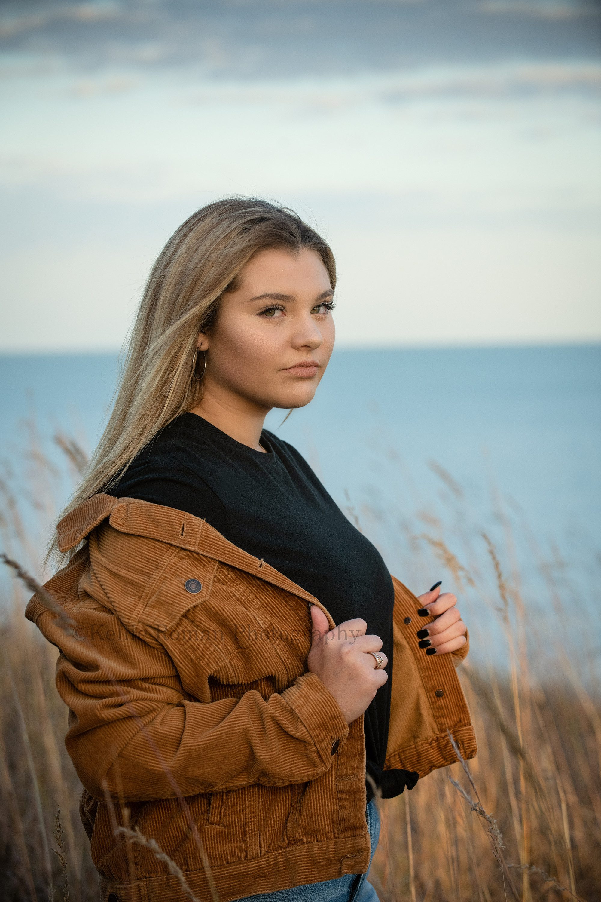 high school senior photographer a high school girl is standing in a milwaukee county park wearing a brown corduroy jacket with a black sweater underneath she is standing on a bluff over looking Lake Michigan and is surrounded by tall grass