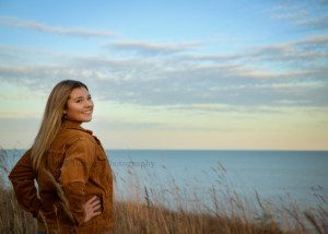 high school senior photographer a high school senior girl is standing in a park along Lake Michigan in Milwaukee county Wisconsin the girl is wearing a brown corduroy jacket and is standing with her hands on her hips twisting backwards to look at the camera she is on a bluff above Lake Michigan with tall grass surrounding her