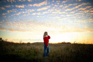 high school senior photographer a senior girl from kenosha is standing in a park in milwaukee county the park is a giant field of tall grass and the sun is setting behind her and the sky is glowing yellow with blues and little white clouds the senior girl is wearing a red sweater and jeans and has blonde hair