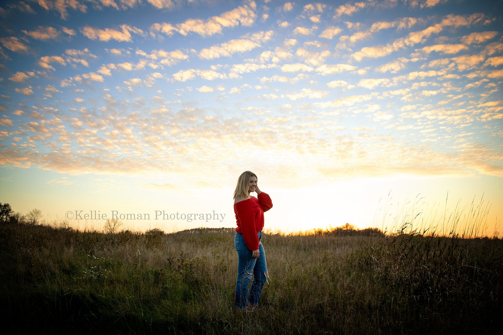 high school senior photographer a senior girl from kenosha is standing in a park in milwaukee county the park is a giant field of tall grass and the sun is setting behind her and the sky is glowing yellow with blues and little white clouds the senior girl is wearing a red sweater and jeans and has blonde hair