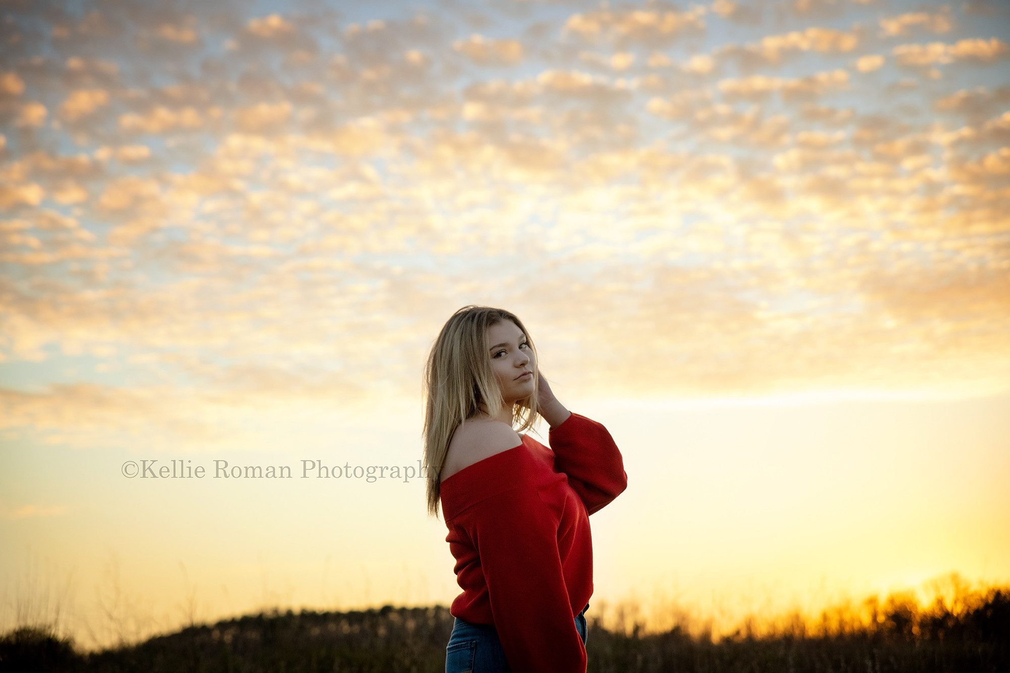 high school senior photographer a senior girl is standing in a field at sun set the sky is bright yellow with blue and small puffy clouds are everywhere the girl is wearing jeans and a red sweater and has blonde hair she has her left arm in her hair and is looking at the camera