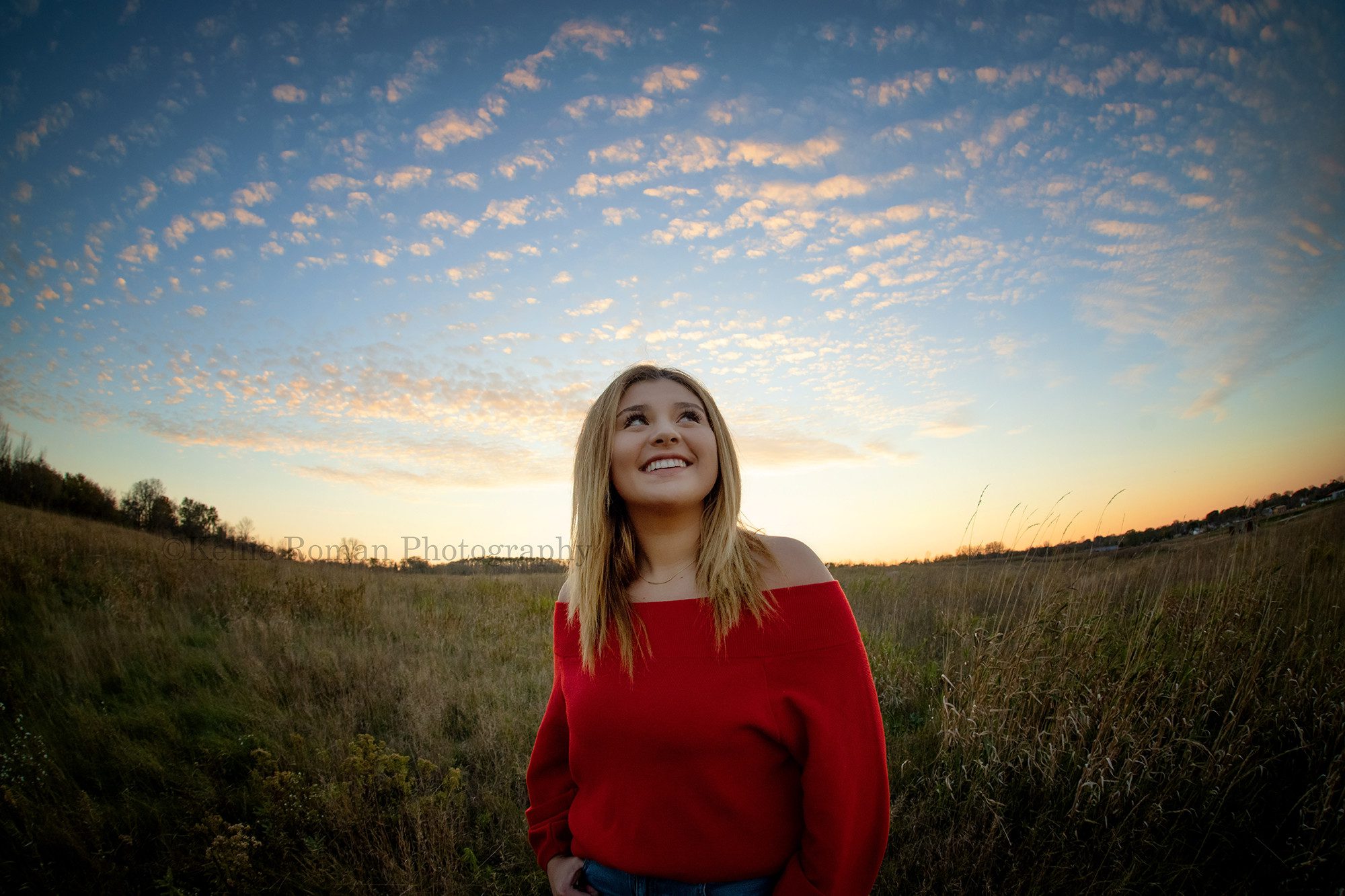 high school senior photographer a high school senior girl is standing in a field of tall grass in milwaukee county she's wearing a red sweater and is looking up towards the sky that is filled with little puffy clouds and beautiful colors