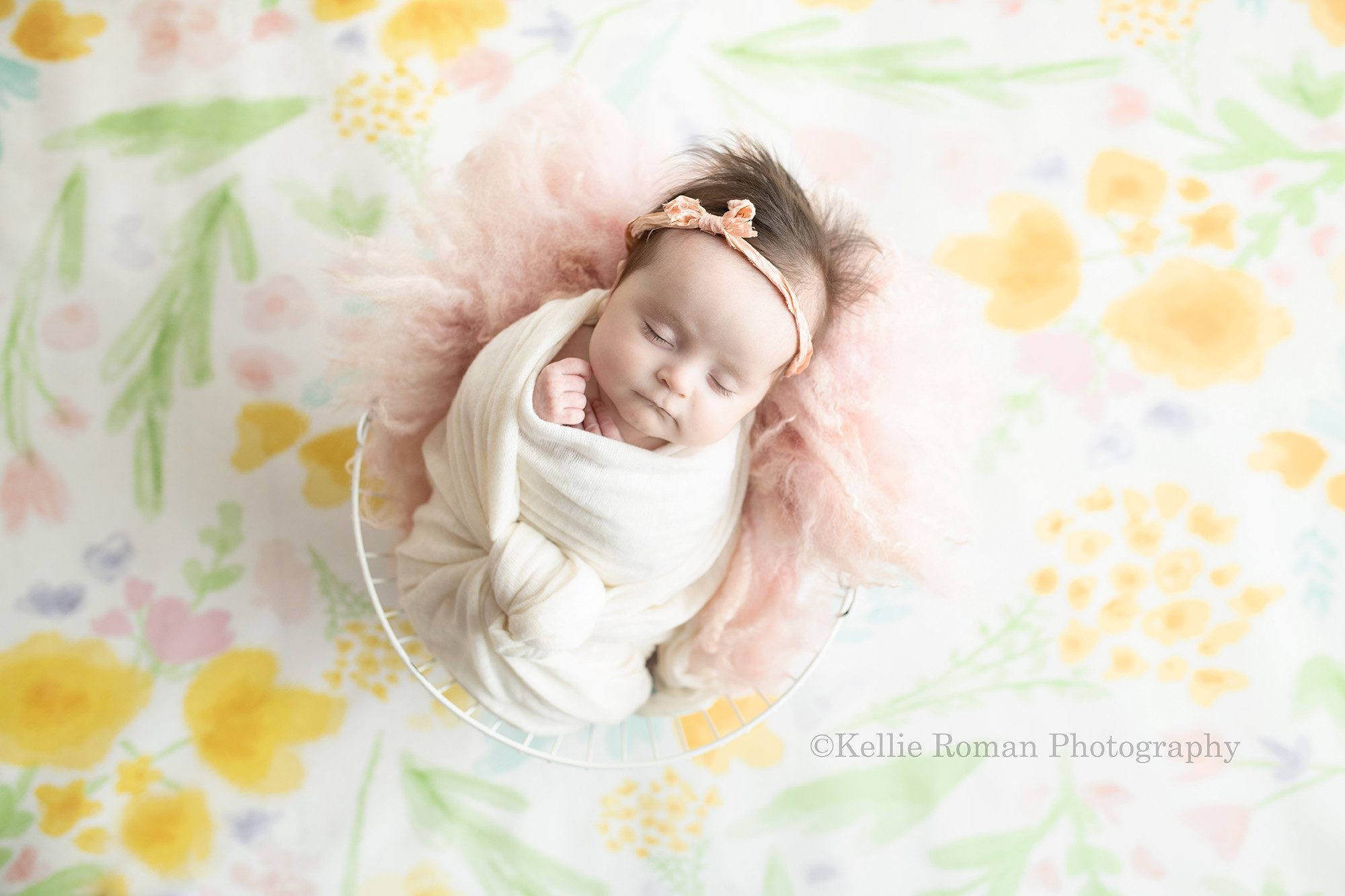 sweet newborn a image of a newborn baby girl in a milwaukee photographers studio she is wrapped in a cream swaddle and she has a light pink bow headband on she is sleeping and is resting on top of light pink fur in a white wire bucket. the bucket is on top of a floral backdrop with shades of pink blue purple and green