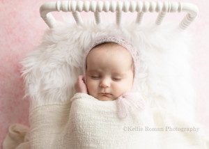 sweet newborn a close up shot of a newborn baby girl in a milwaukee photographers studio. The studio is in greendale Wisconsin. The baby girl has on a light pink mohair bonnet and is laying on top of white fur on a white wood bed. Her right hand is up by her cheek and she's sleeping. The bed is on top of a light pink backdrop.