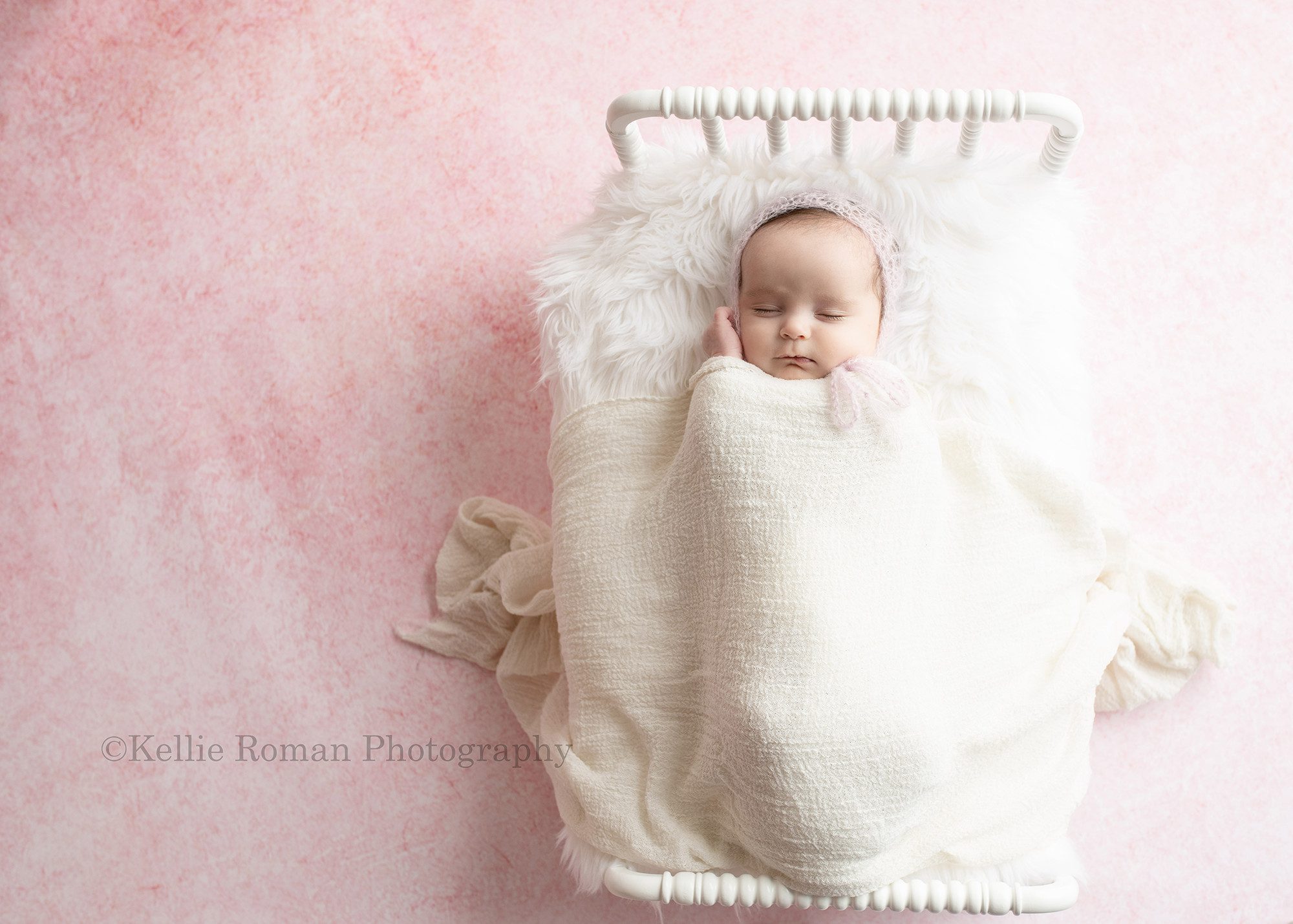 sweet newborn a baby girl is in a milwaukee Wisconsin photography studio in greendale. The baby is sleeping on a white wood bed and has a light pink bonnet on with a white wrap covering her the bed is on top of a light pink backdrop