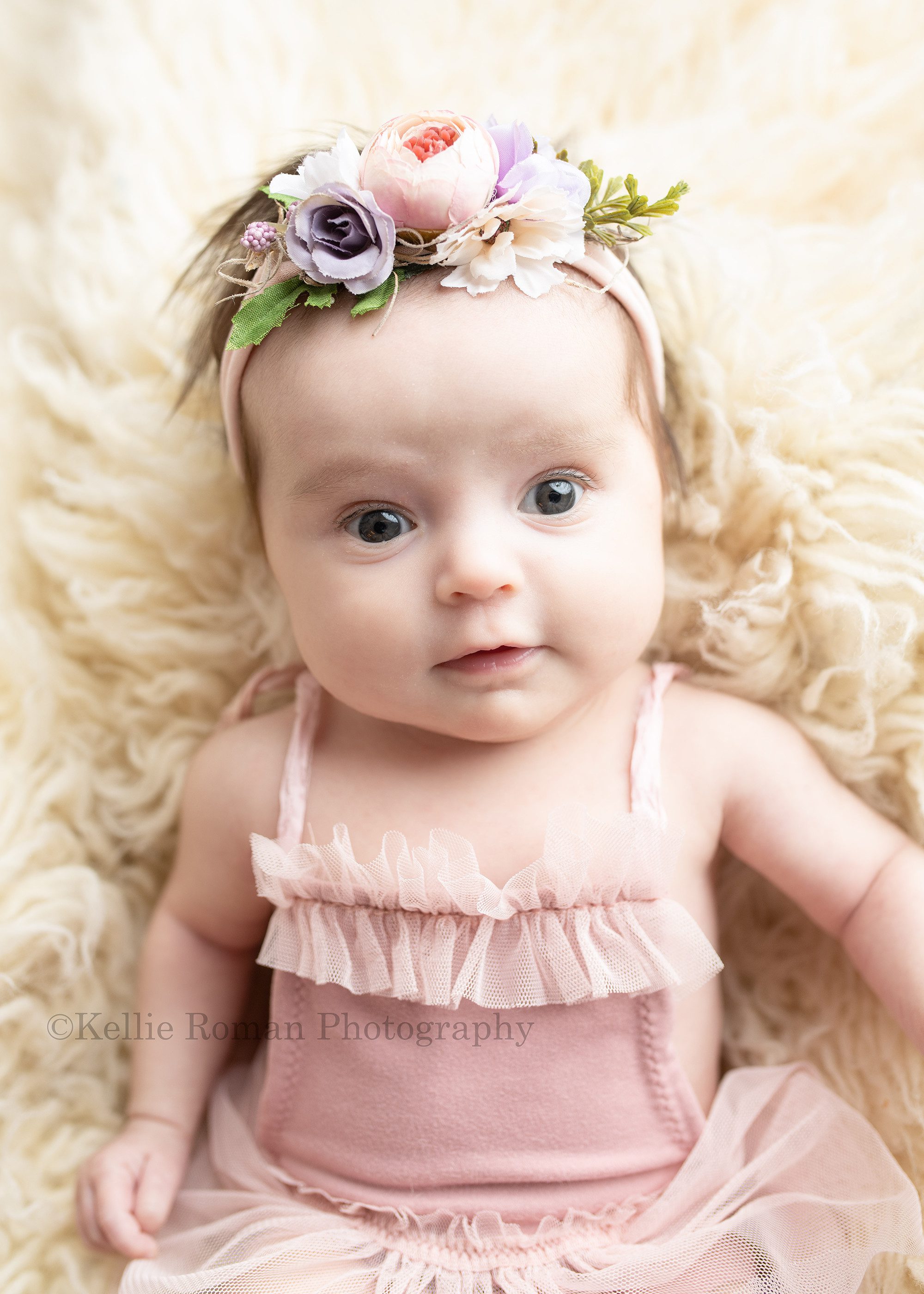 sweet newborn a newborn baby girl is in milwaukee photographers studio she is laying on top of cream color fur rug and is wearing a blush colored tutu newborn romper and a blush and purple floral headband she has a serious face but is looking into the camera