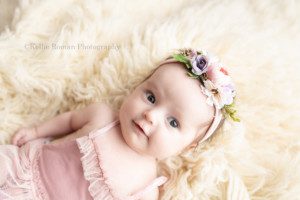 sweet newborn a two month old baby girl is in a greendale photographers studio she is laying on her back on top of cream color fur the baby is wearing a blush pink tutu romper and a pink and purple headband the baby girl is smiling and looking off to the side