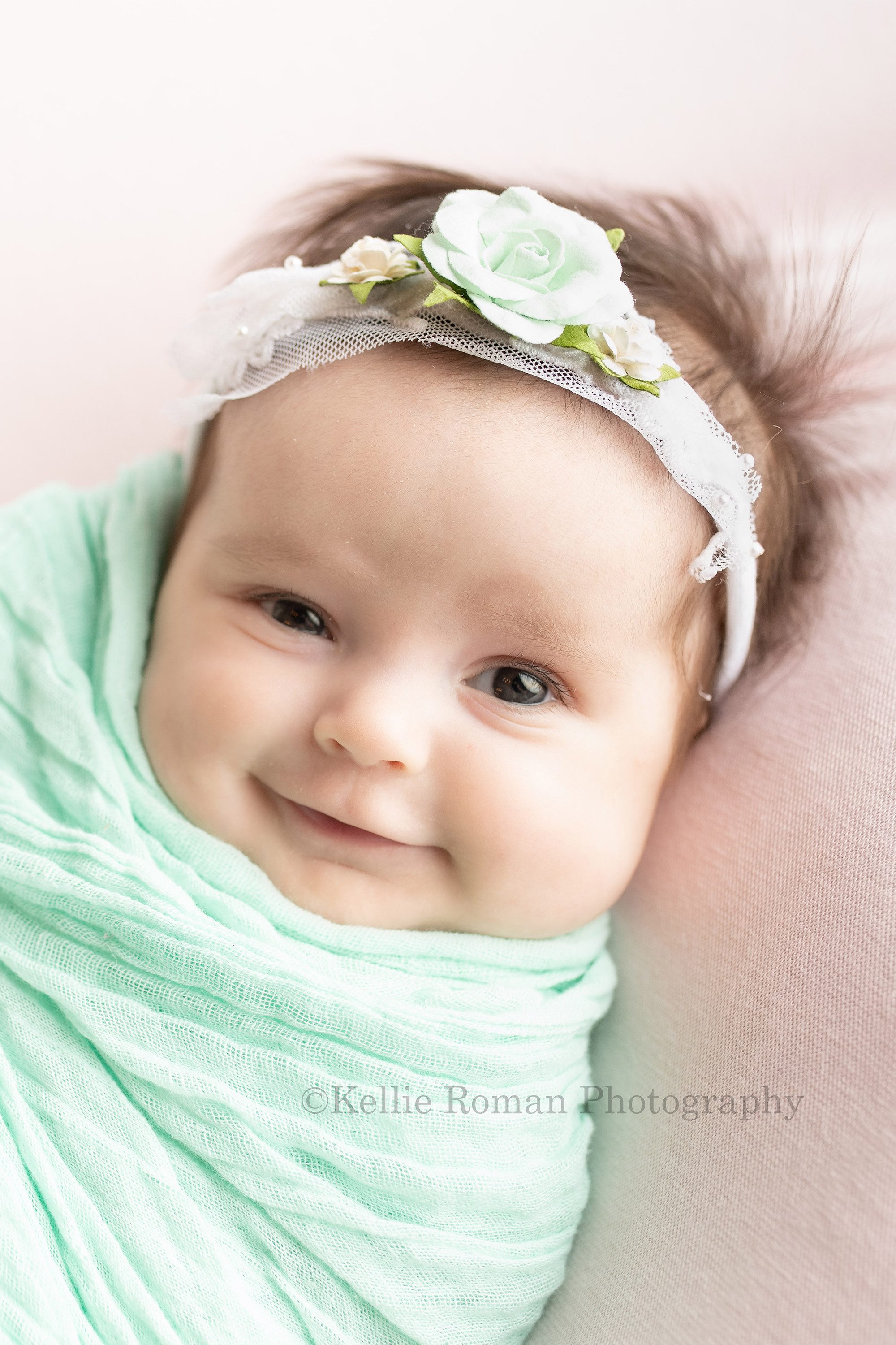 sweet newborn a baby girl in a milwaukee photography studio is posed in a mint color swaddle wrap laying on her back on top of light pink fabric she has a white and mint flower headband, the baby is looking into the camera and smiling