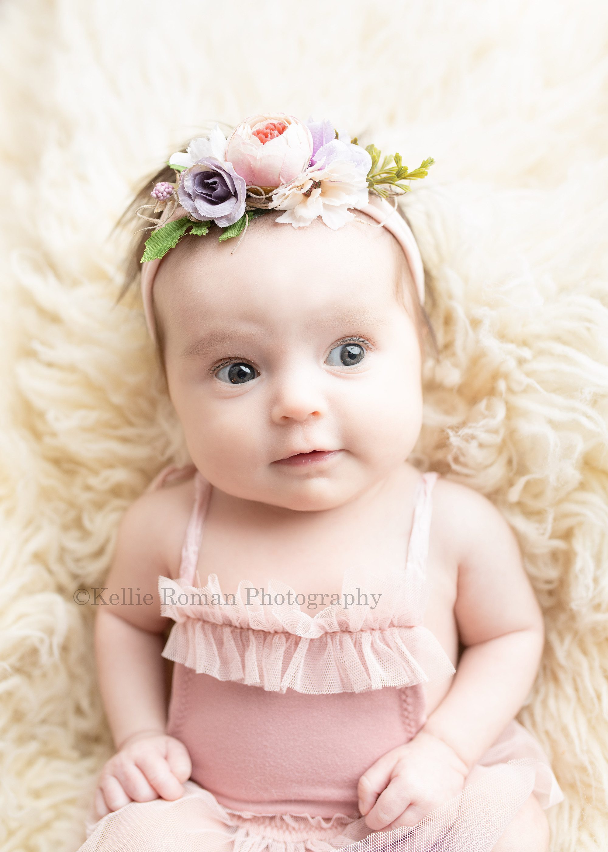sweet newborn a newborn baby girl in a milwaukee photographer studio she is laying on her back on top of cream colored fur she has on a blush color tutu romper and a pink and purple floral headband she is looking up and off to the side and her arms are down at her side