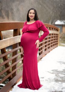 maternity in the snow a women in a dark red dress is standing in pure white snow on a bridge in a milwaukee county park. She has her left hand on her hip and is looking and smiling into the camera.