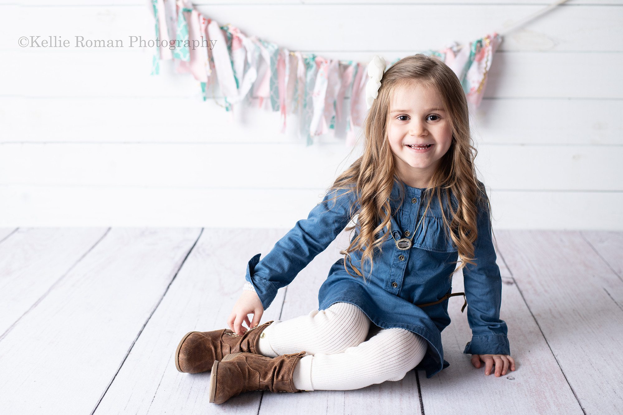 six month milestone a young girl is in a milwaukee photo studio having her pics taken. she is wearing a denim dress with white leggings and brown boots. She has long dark blonde hair and is smiling at the camera. she's sitting on a white wood floor in front of a white wood backdrop and it has a teal and pink fabric banner hanging.
