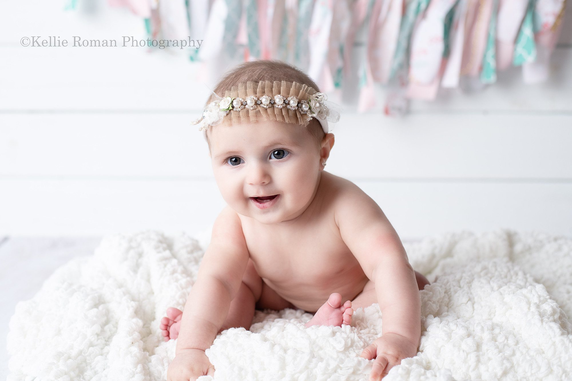 six month milestone a baby girl is smiling while having her pics taken in a Milwaukee photographers studio located in greendale Wisconsin. The girl is naked and sitting on a white fluffy blanket in front of a white wood backdrop and pink banner. She has a girlie headband on.