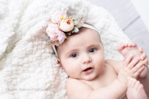 six month milestone a baby girl is laying on her back onto of a white blanket playing with her toes. She is in a milwaukee photo studio located in greendale Wisconsin. The baby girl is smiling and has a big flower headband on.