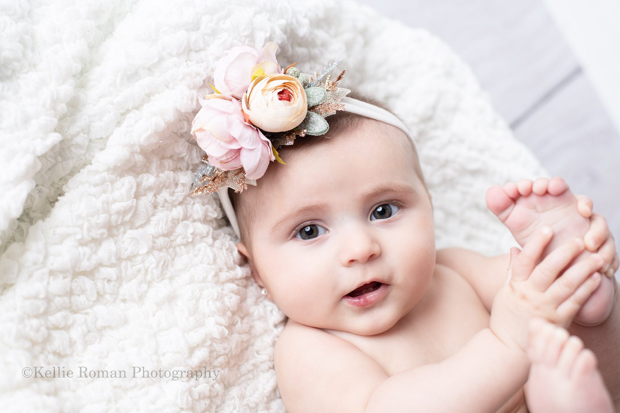 six month milestone a baby girl is laying on her back onto of a white blanket playing with her toes. She is in a milwaukee photo studio located in greendale Wisconsin. The baby girl is smiling and has a big flower headband on.