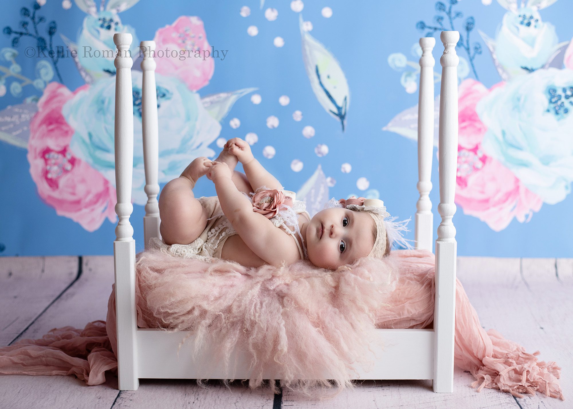 six month session a sweet baby who is six months old is being photographed in milwaukee studio she's on a white four post bed on top of pink fluff and fabric. she's playing with her toes on her back, and the backdrop is blue with white and pink florwers.