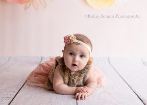 six month milestone a baby girl is in a milwaukee photography studio she is wearing a brown fur vest and a peach skirt she's laying on her bellow on a white wood floor and in front of a peach colored backdrop the girl has her hands on top of each other