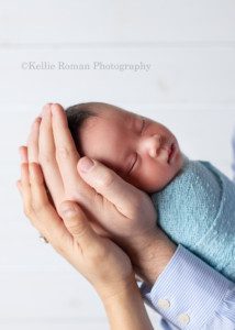 bright airy newborn session. A newborn baby boy is in a milwaukee photography studio. The baby is sleeping and wrapped in a blue swaddle. Dad has his sons head in his hands, and moms hand is resting on dads.