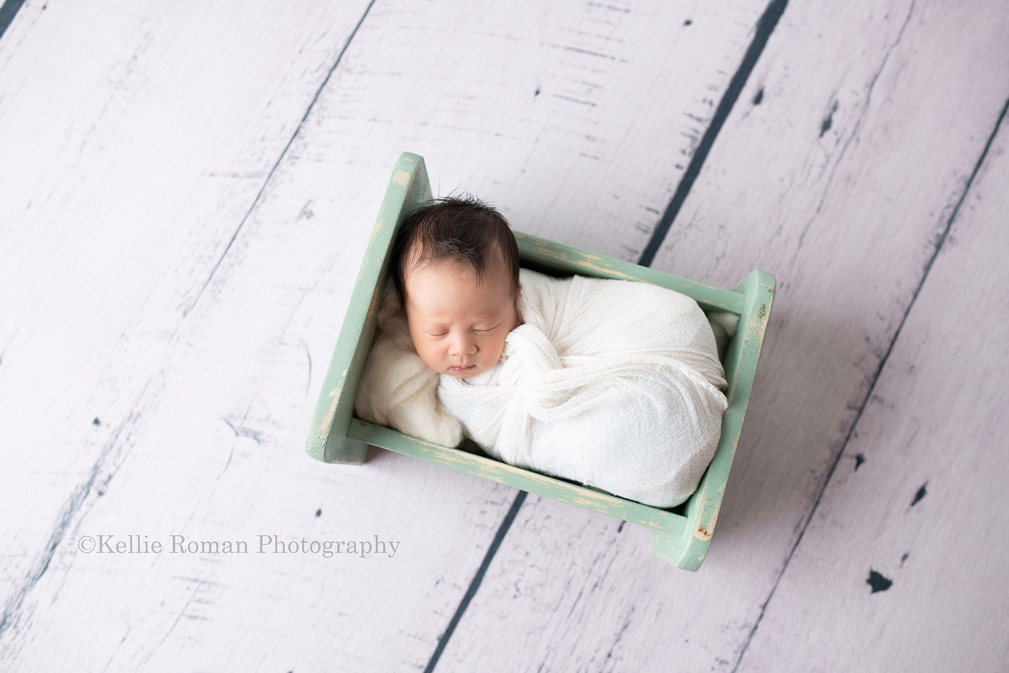 bright airy newborn session. A newborn baby boy is in a milwaukee photo studio. The baby is wrapped in a white swaddle wrap and is wearing a white sleepy hat with black stars. The baby is resting on white fluff and is sleeping on his back on a teal bed.