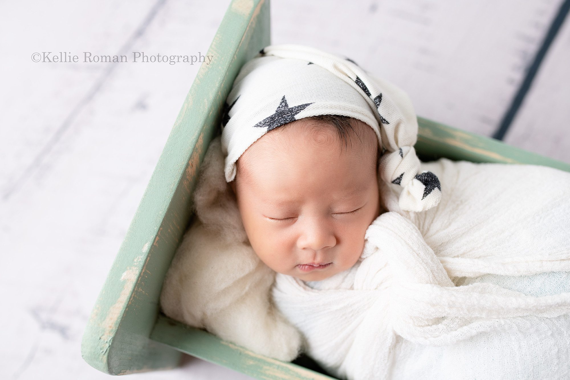 bright airy newborn session a newborn baby boy in a milwaukee photography studio located in greendale Wisconsin. The baby boy has a white swaddle wrap around him and a white hat with black stars on. He is resting on his back asleep on white fluff in a teal bed.