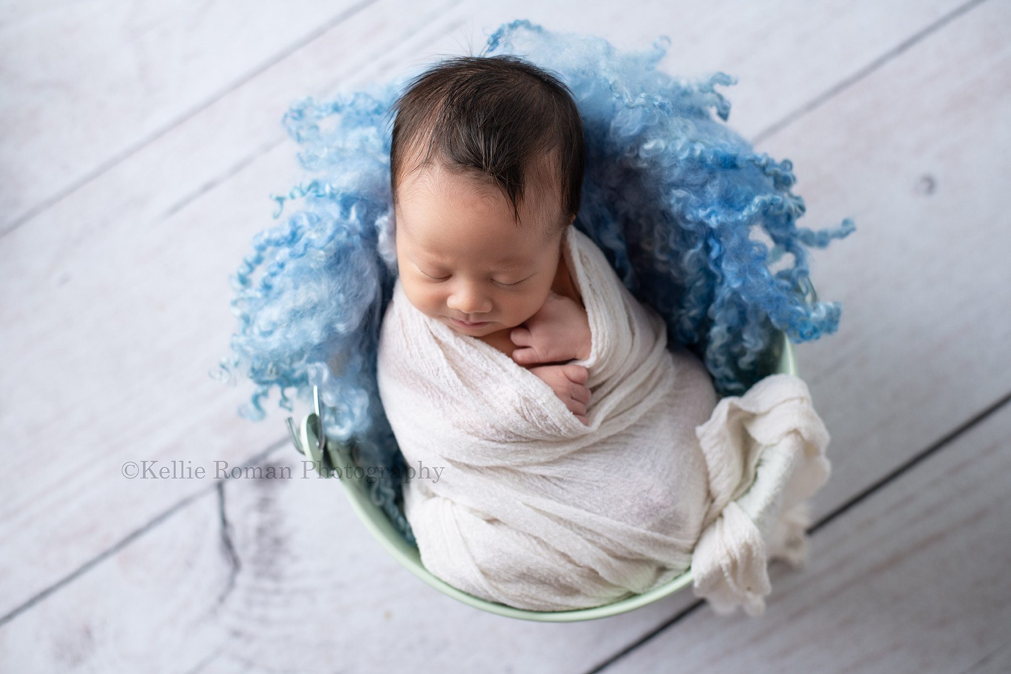 bright airy newborn session a little baby boy in a milwaukee photo studio is swaddled in a white wrap he is resting on his back in a metal mint bucket with blue fur the bucket is on top of a white floor. The baby has his hands resting on his chest.