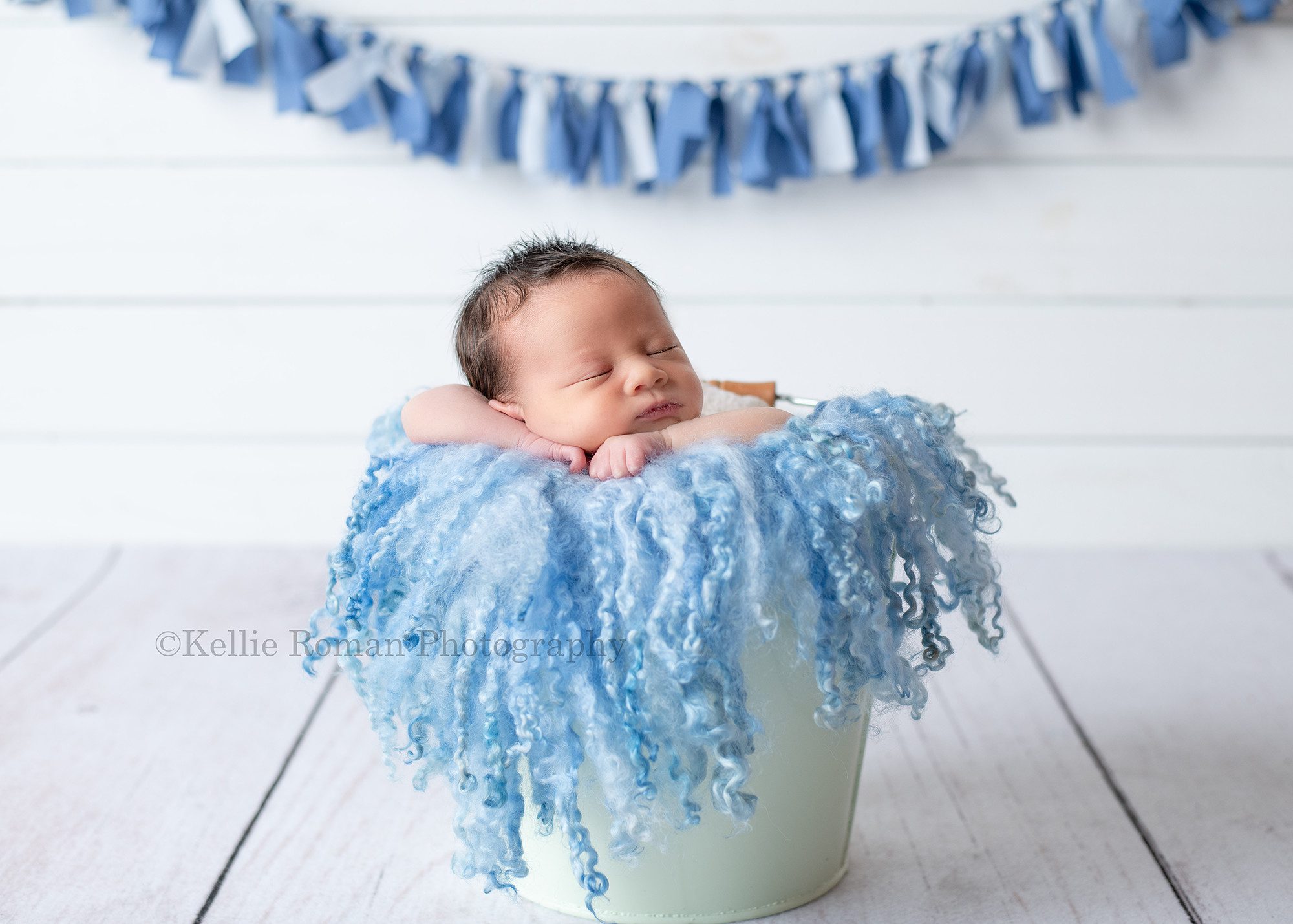 bright and airy newborn a baby boy in milwaukee photographers studio the baby is posed upright in a mint metal bucket with a blue curly wool layer in it the bucket is onto of white wood flooring and in front of a white wood wall with a blue fabric banner behind