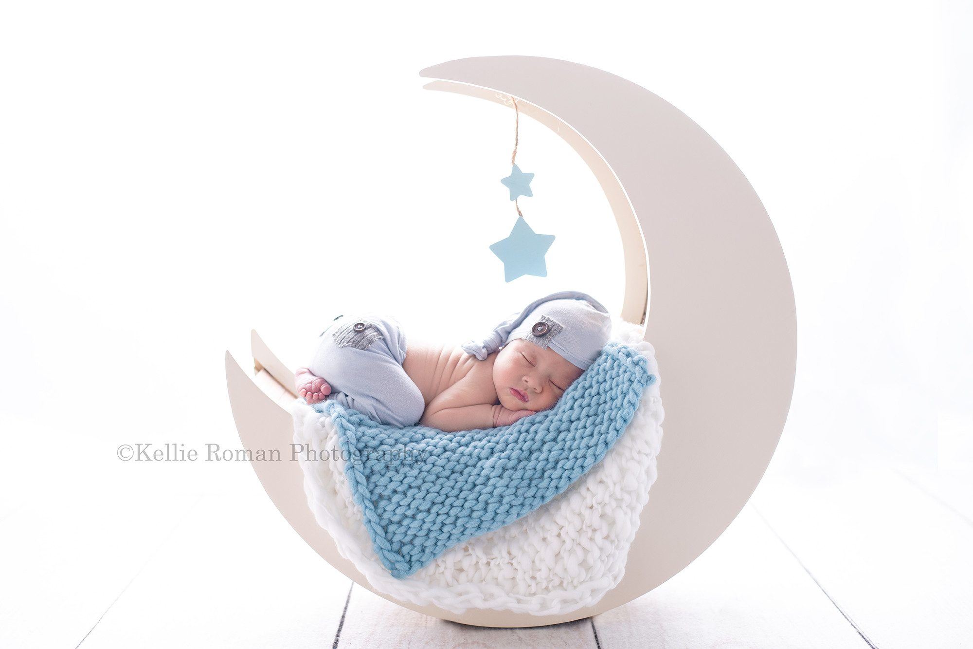 bright airy newborn session a newborn baby boy is in a milwaukee photography studio. He is posed on his belly sleeping on a big white wood moon prop. There are blue and white layers of fabric on the prop. and the boy is wearing blue pants with a matching blue hat. The backdrop is very bright and white.