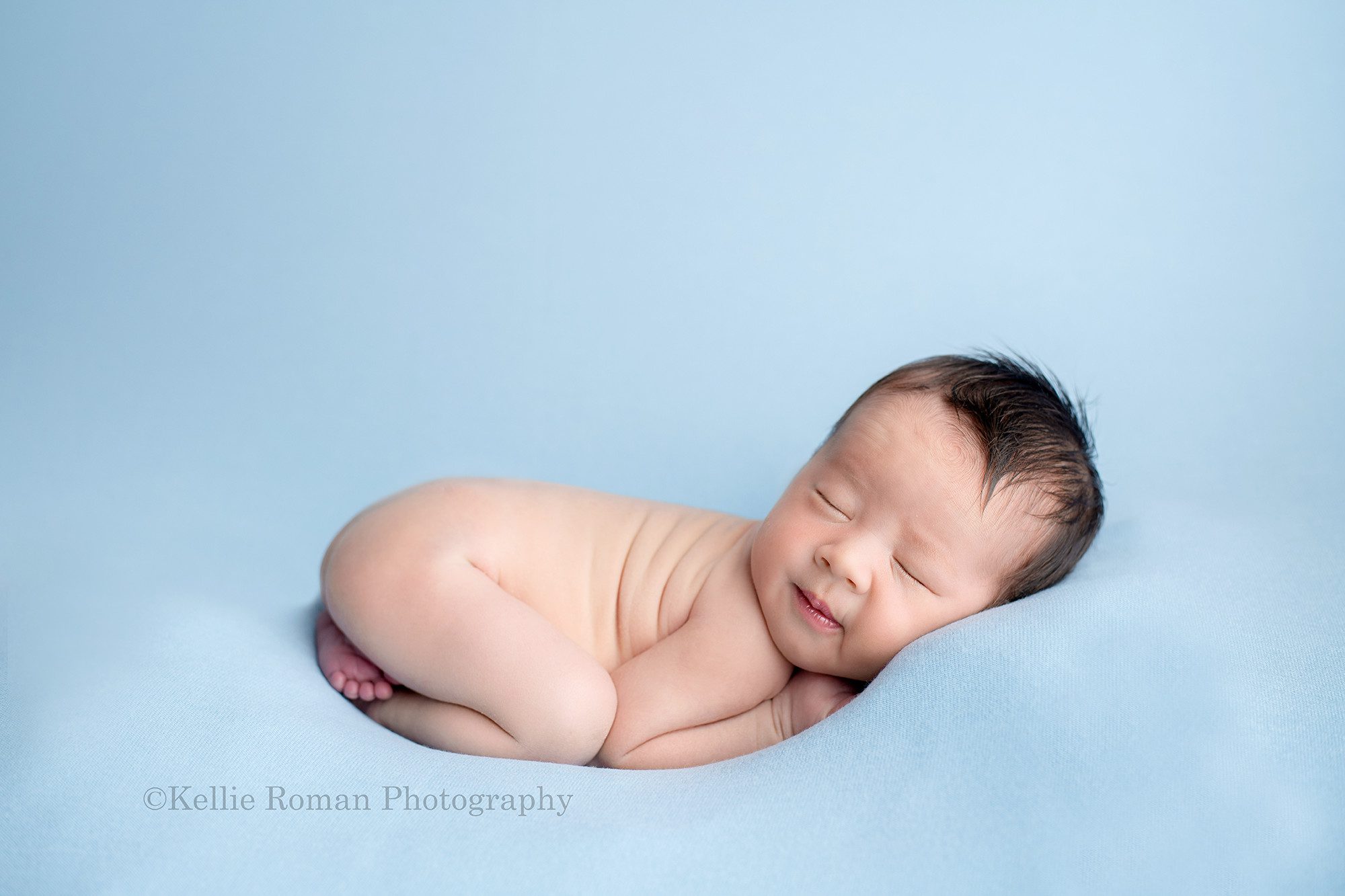 bright airy newborn session a little boy in a milwaukee Wisconsin photographers studio he is posed on his belly curled up onto of a blue blanket the little boy has brown hair and is smiling in the shot