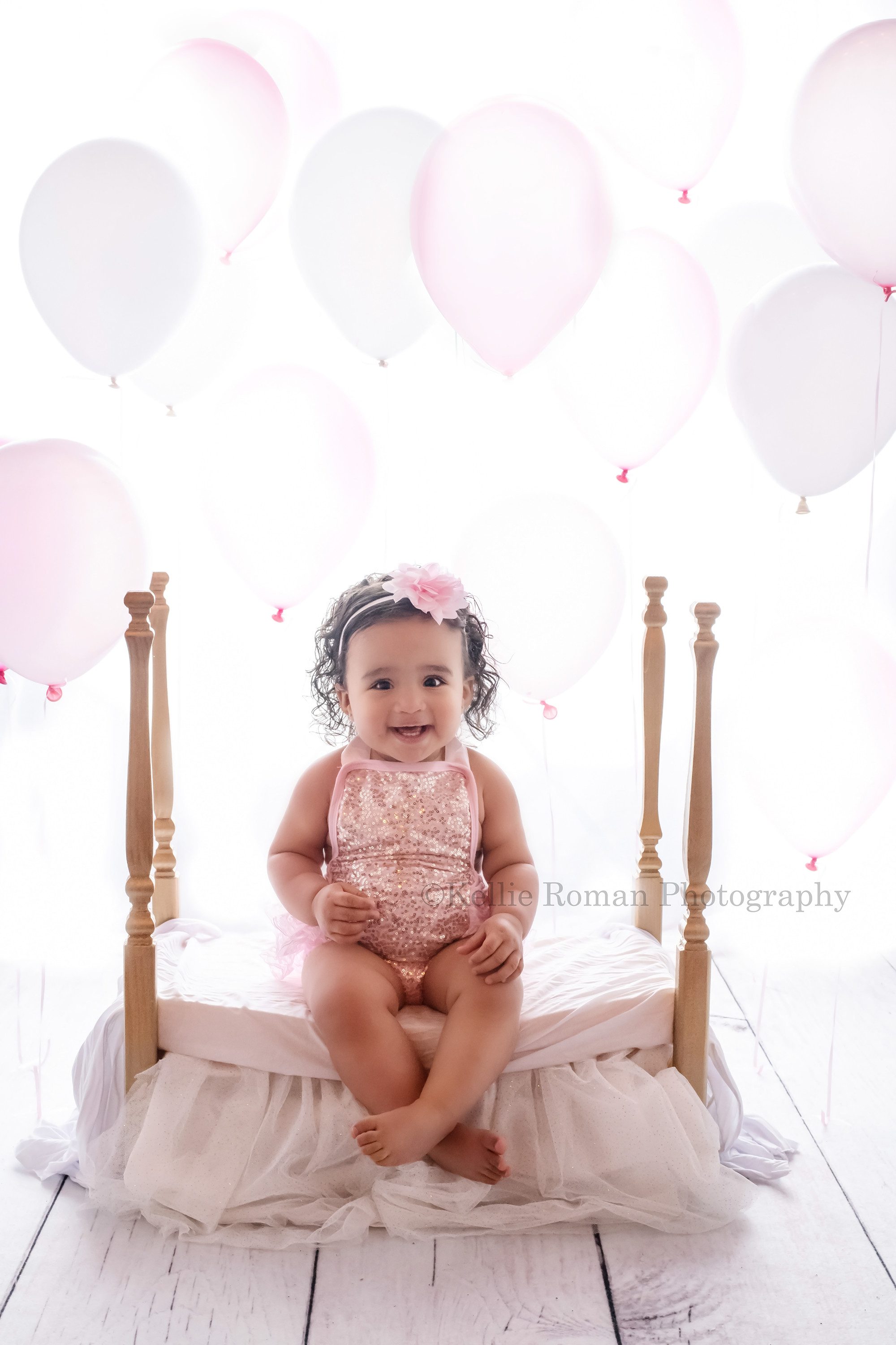 milwaukee cake smash photographer a one year old little girl is in a milwaukee Wisconsin photography studio in greendale. She is wearing a pink and gold sequin romper and is sitting on a gold four post bed she's smiling at the camera and crossing her ankles. The backdrop is bright white with pink and white balloons.