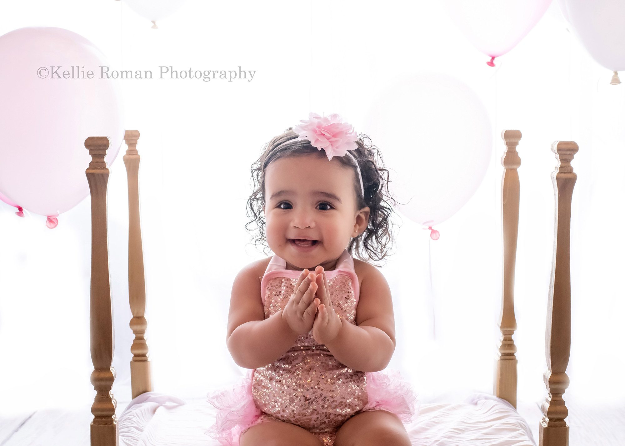 milwaukee cake smash photographer a one year old girl with brown curly hair is in a milwaukee photography studio located in greendale Wisconsin she's wearing a pink and gold sequin romper and is sitting on a gold four post bed she's clapping and smiling and has pink and white balloons behind her