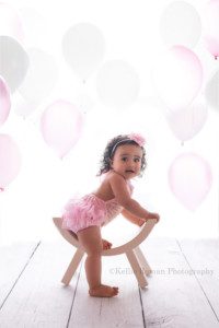 milwaukee cake smash photographer a one year old girl in a milwaukee photographers studio in greendale Wisconsin she is sitting on a white curved bench with one leg down on the floor the backdrop is bright white with pink and white balloons behind her the girl has brown curly hair and is wearing a pink and gold romper