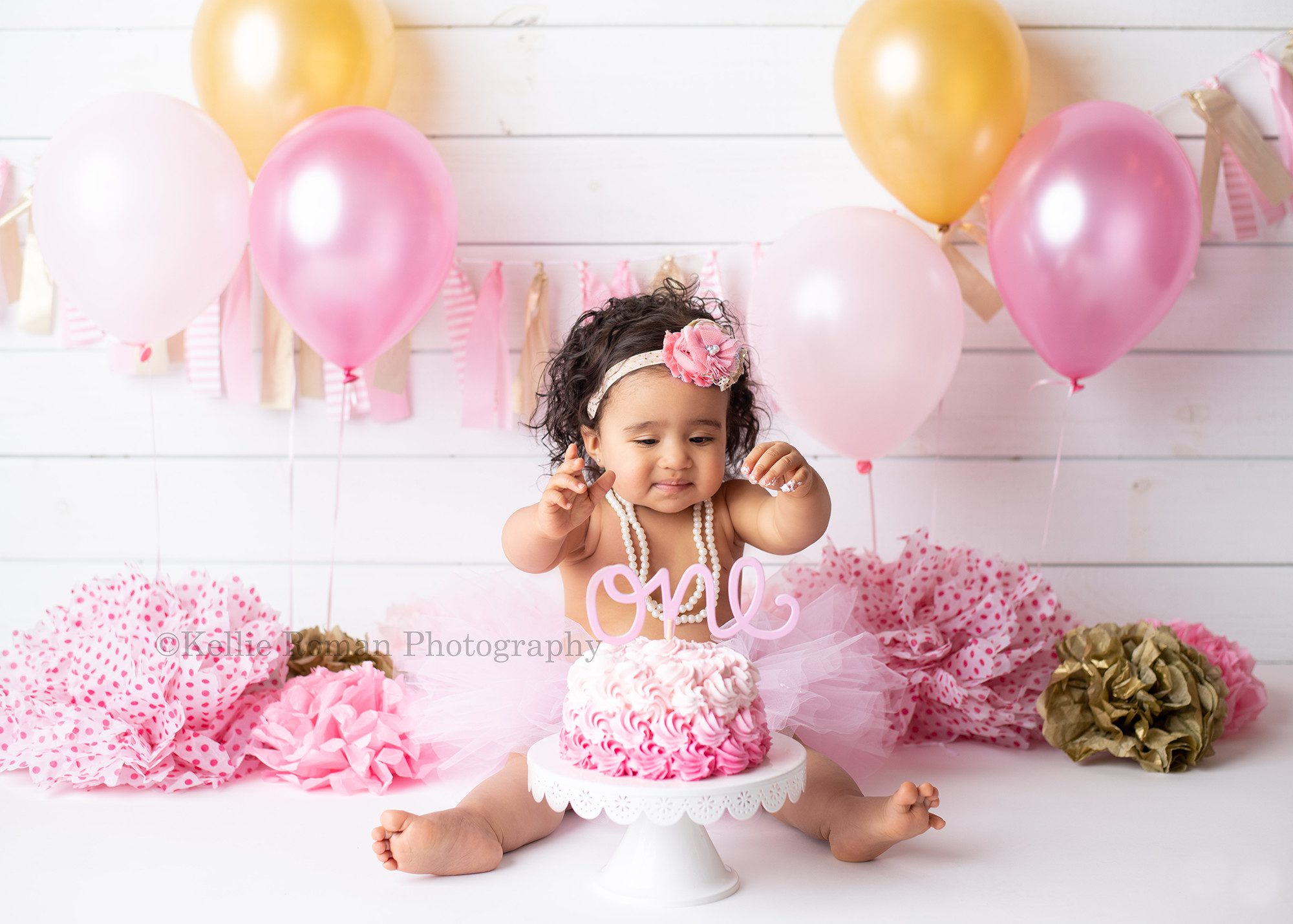 milwaukee cake smash photographer a one year old little girl in a milwaukee greendale photography studio sitting behind a ombre pink swirl cake on a white cake stand she's wearing a pink tutu with pearl necklaces and has her hands in the air she has pink pom poms behind her on the floor with gold and pink balloons and a gold and pink banner