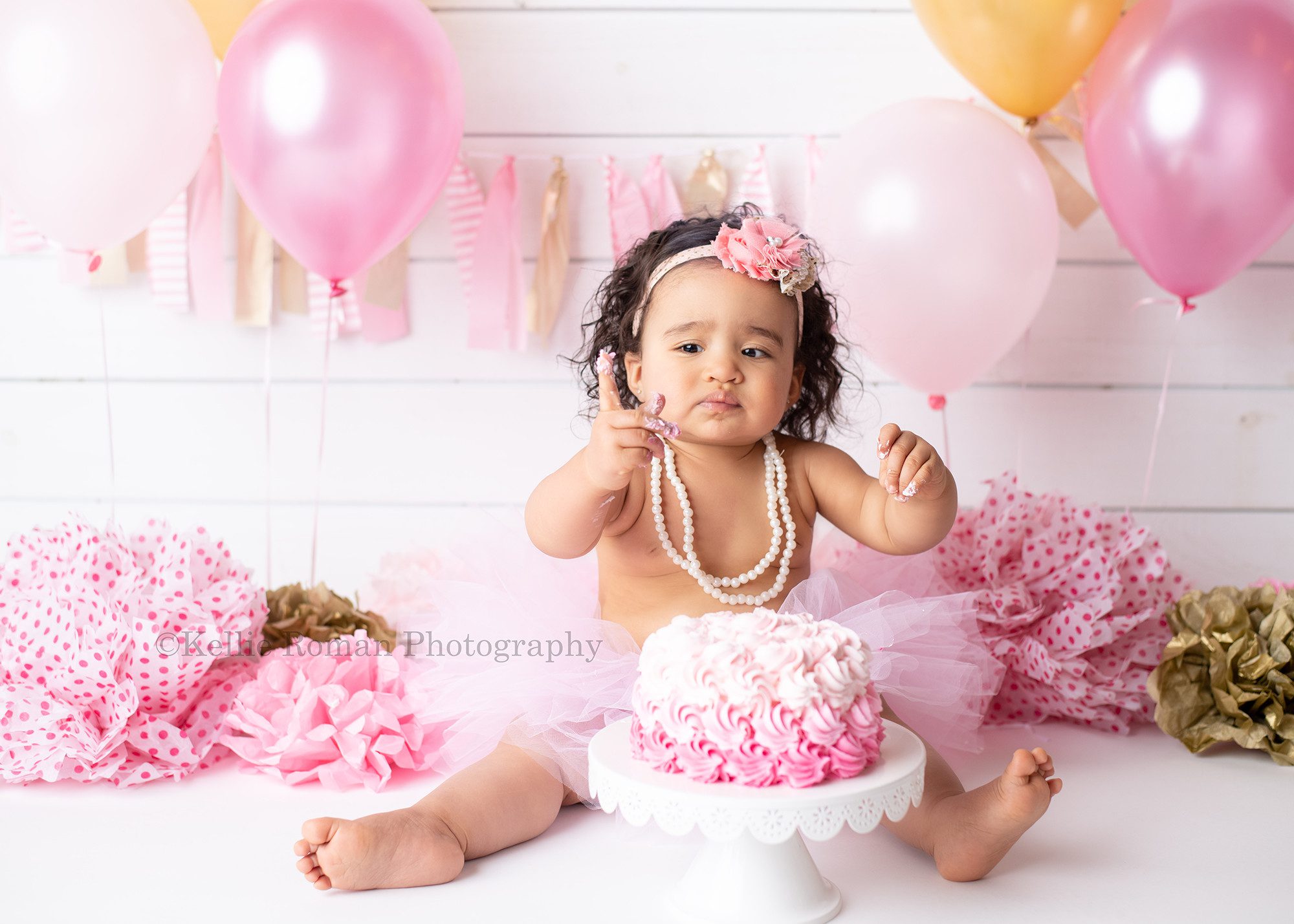 milwaukee cake smash photographer a one year old girl is having her milestone session in a milwaukee photo studio in greendale Wisconsin. The girl is wearing a pink tutu with pearls and is sitting behind a pink swirl cake thats resting on a white cake stand there are pink and gold balloons pink pom poms and a pink and gold banner behind her