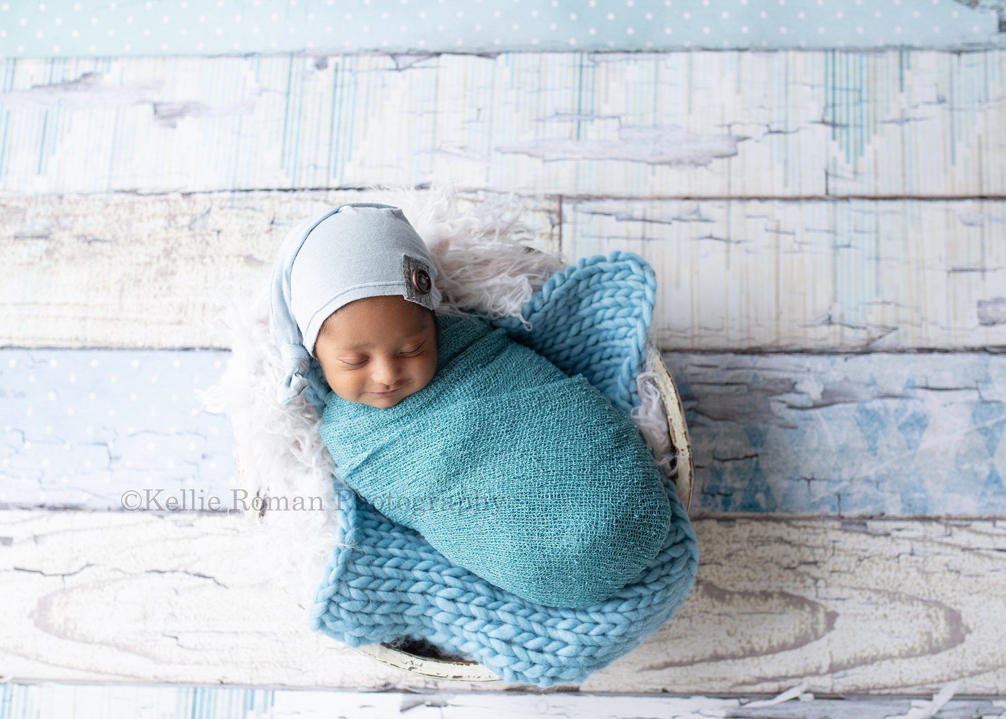 sweet baby boy. A newborn boy in a milwaukee photographer studio is wrapped in a blue swaddle. He's laying in a white wood tub filled with cream fur and a blue fabric blanket. He has on a hat with a brown button and he is smiling in his sleep.