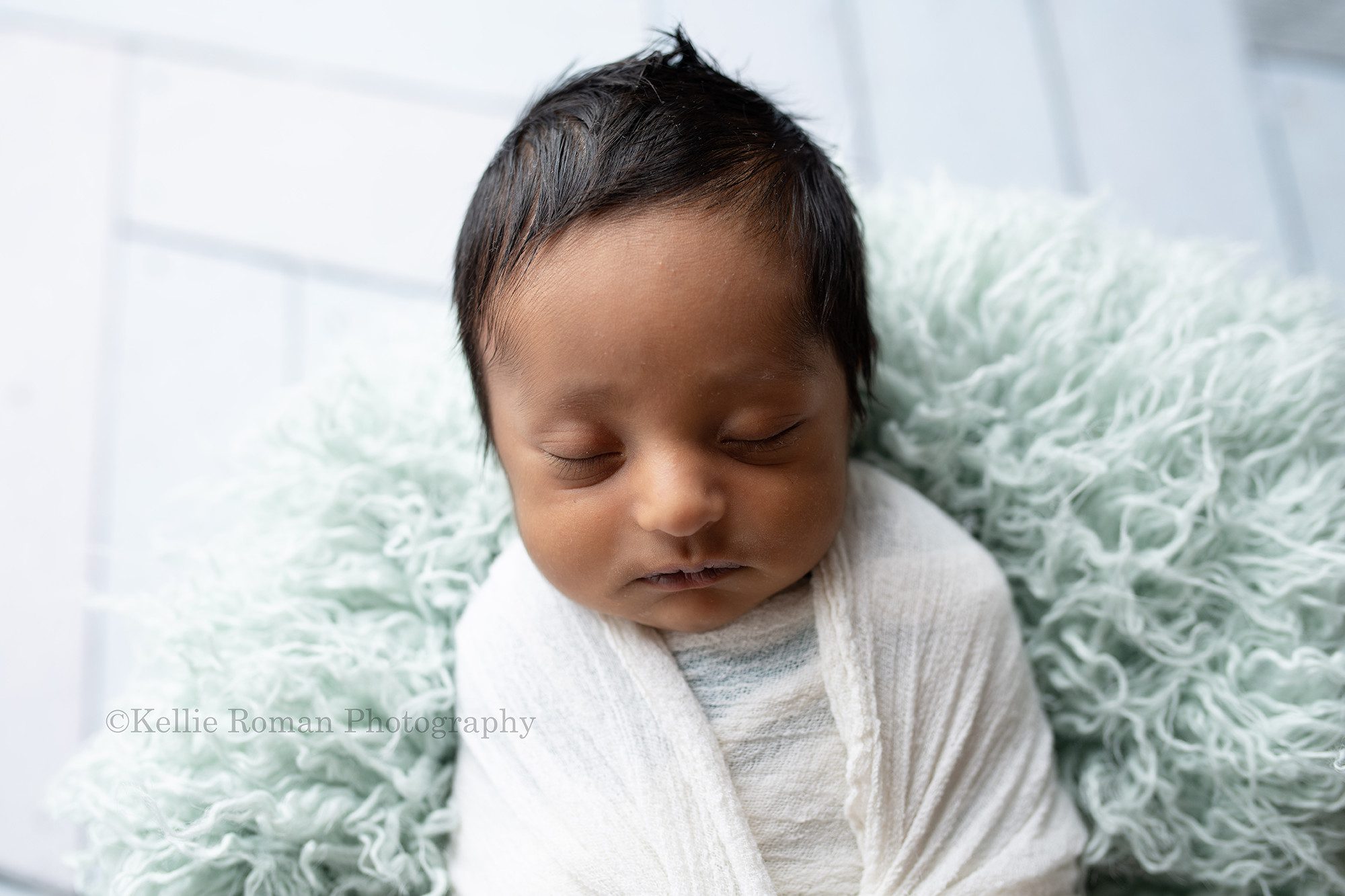 sweet baby boy a newborn little boy in a greendale Wisconsin photographers studio he is wrapped in a cream colored swaddle fabric. He's laying on his back in a white metal basket with teal fur underneath him. He has lots of very dark hair.