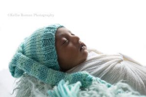sweet baby boy a newborn boy with lots of dark brown hair has a teal colored sleepy cap on. He's wrapped in a white swaddle fabric and is laying on his back sleeping in a metal bucket with fur. The lighting is coming from behind him and is very bright. The shot is a profile image of his face.