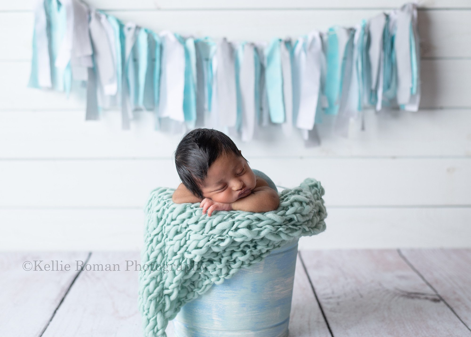 sweet baby boy a baby boy in a milwaukee photography studio. The baby boy has lots of dark hair and is proped upright in a blue metal bucket with teal blanket the babies chin is resting on his arms. There is a white wood wall behind him with a blue fabric banner.
