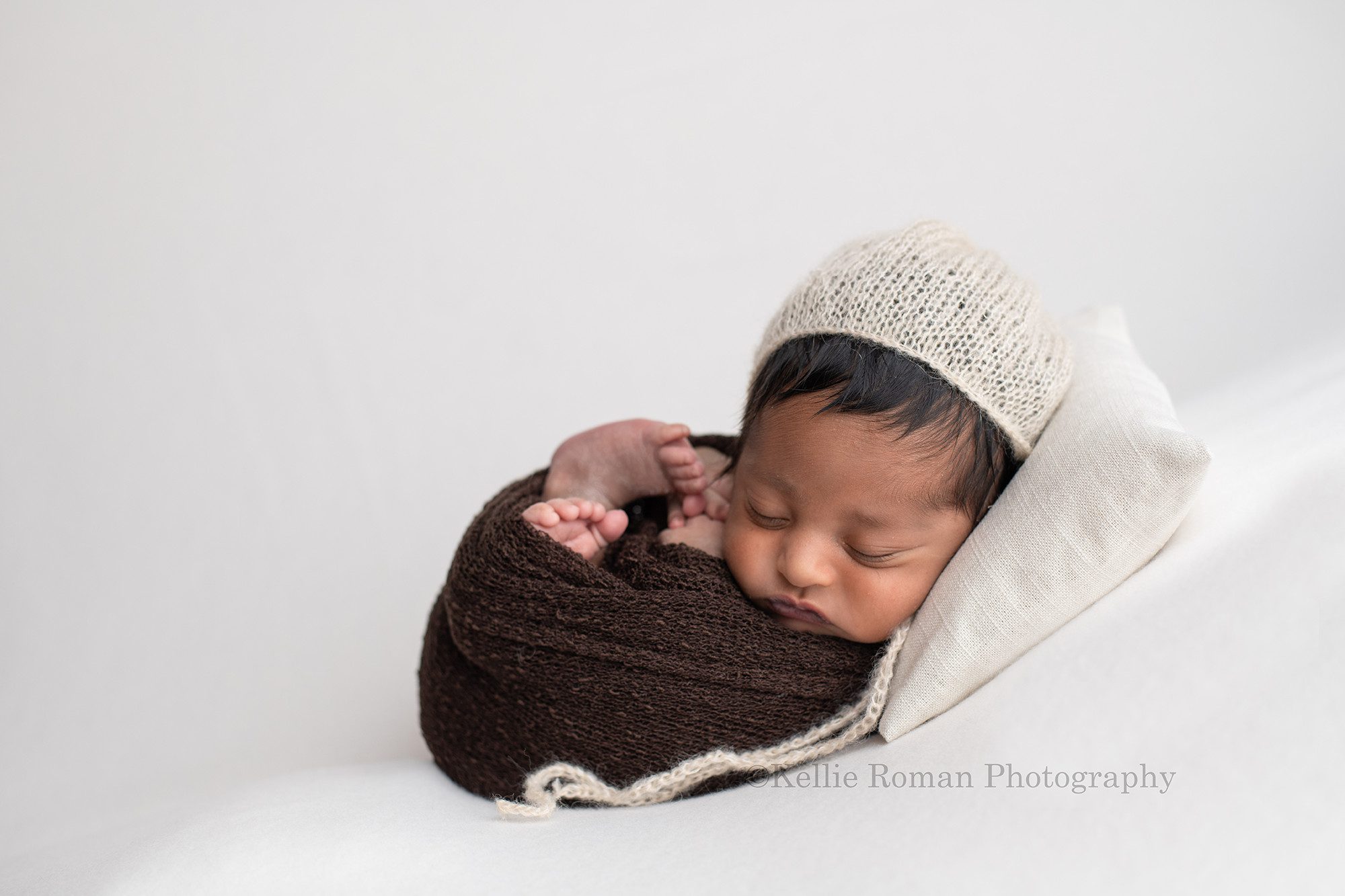 sweet baby boy a newborn baby boy in a milwaukee photographers studio he is wrapped in a brown swaddle fabric and is laying on his back on a cream color fabric. He has a cream color bonnet on, and is head is resting on a cream colored pillow.