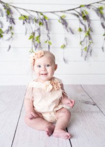 happiest six month old. A baby girl in a greendale photographers studio is sitting on white wood flooring with a white wood wall behind her. She has an ivory romper on with feathers and flowers and a matching headband. She's looking into the camera with big blue eyes.