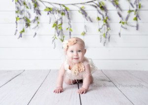 happiest six month old a young baby girl in a milwaukee photographers studio is on her hands and knees with a big smile on her face. She's on a white wood floor with a white wood backdrop and lilac vine behind her. she's wearing pale pink and ivory colors and has a headband in her hair