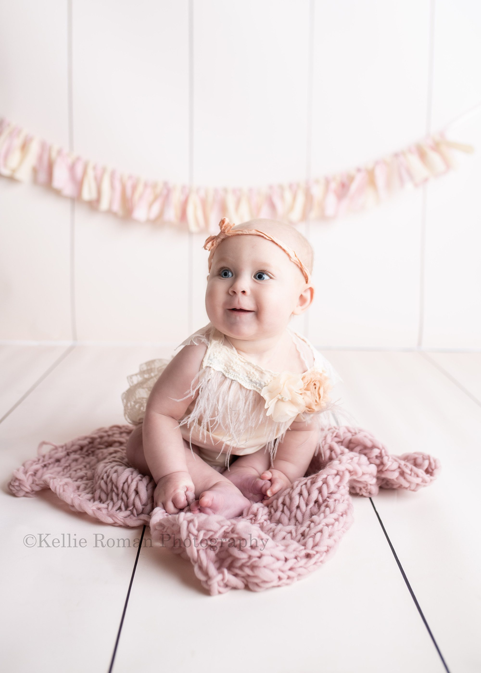 happiest six month old. A baby girl in a greendale studio is sitting on top of blue knit blanket. The floor and backdrop both match and it's ivory wood. She's smiling very big and wearing an ivory romper with a matching headband.