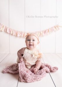 happiest six month old. A baby girl in a greendale photographers studio is siting on a blush colored blanket. She's on top of an ivory floor with a matching ivory backdrop and a fabric banner. The baby is wearing an ivory romper with feathers and a matching headband.