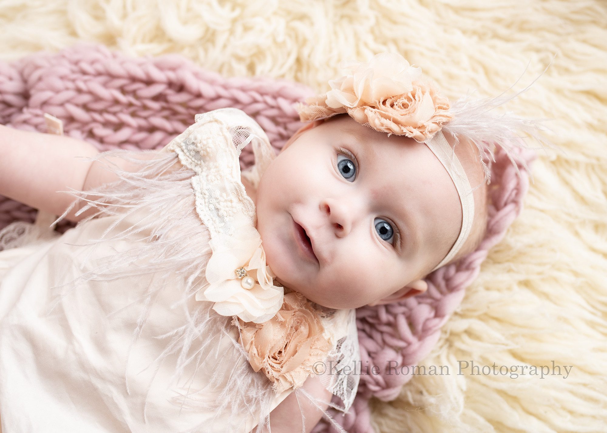 happiest six month old a baby girl is laying on her back in a milwaukee photo studio. She is wearing an ivory romper with feathers and flowers and matching headband. There are layers of fur and fabric under her. she's smiling and looking into the camera.