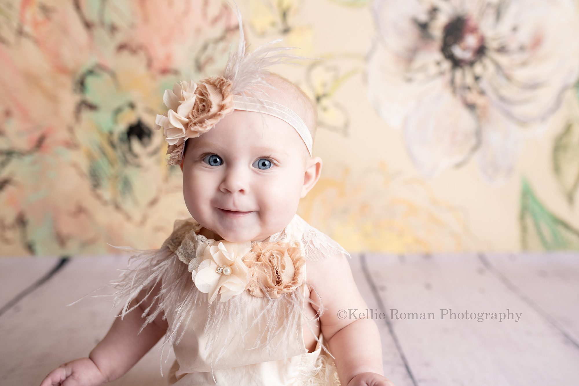happiest six month old. A baby girl wearing a fancy ivory romper with feathers and flowers is in a milwaukee photo studio. She has a matching headband on and is sitting in front of a floral ivory backdrop and wood floor. she's smiling at the camera and has big blue eyes.