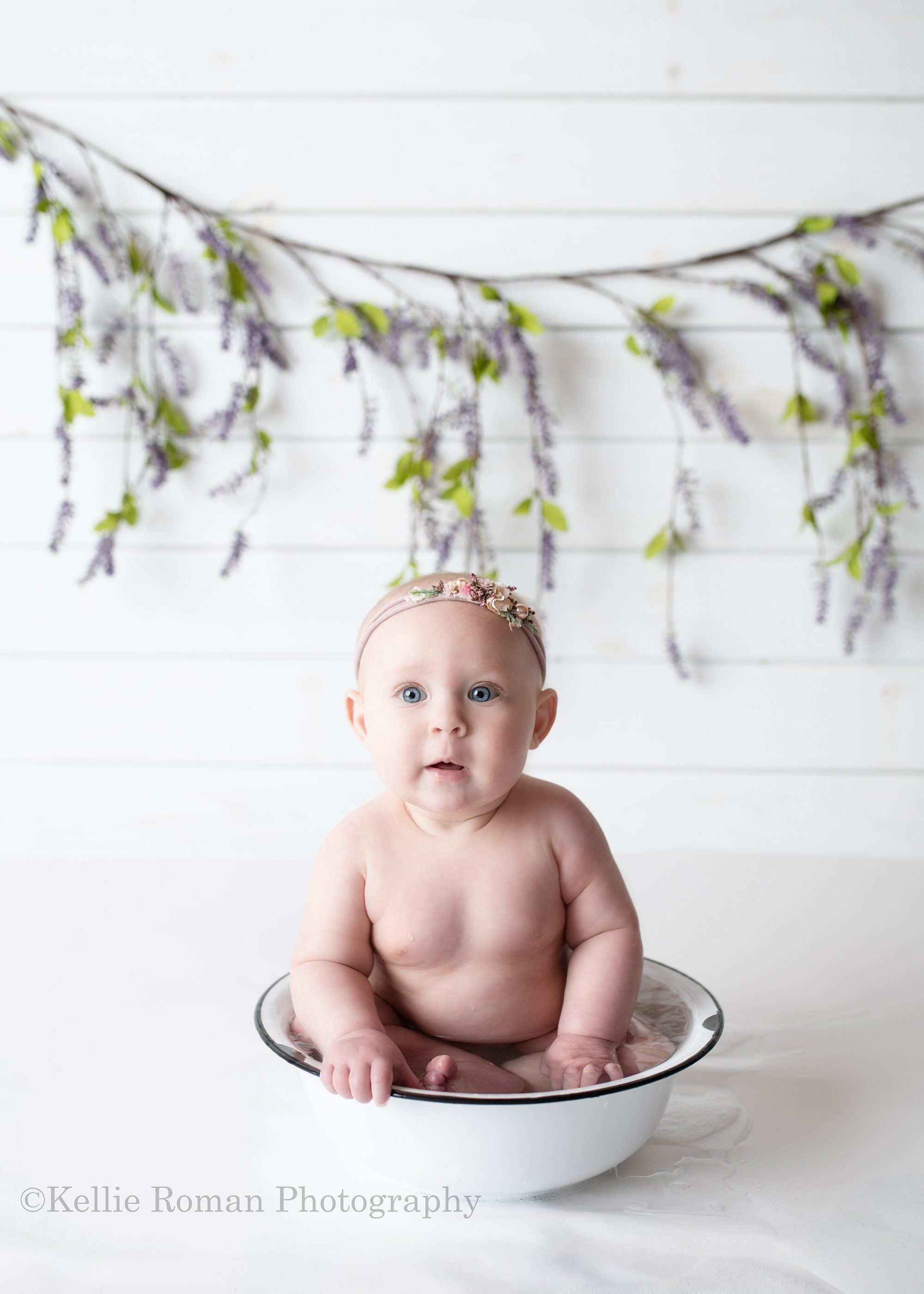happiest six month old. A baby girl is sitting in a white vintage bowl with water and soap. There is water all over the white floor and she is in front of a white wood wall with lilac vine behind her. She has on a light purple floral headband and is looking into the camera with big blue eyes.