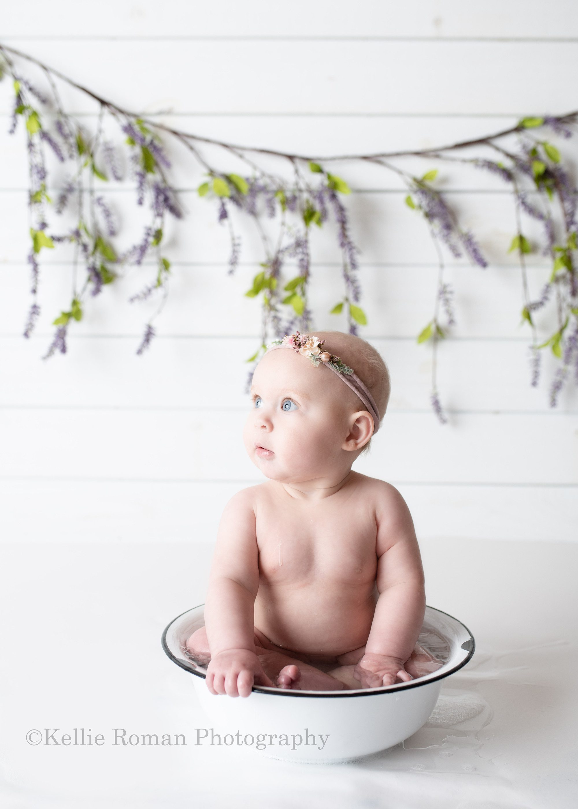 happiest six month old. a six month old baby girl is in a milwaukee Wisconsin photo studio. She's sitting in a white vintage bowl with lots of water in it. The water is all over the white floor. The baby is looking off to the side with a serious look. She has on a headband and is sitting in front of a white wood wall with a lilac vine.