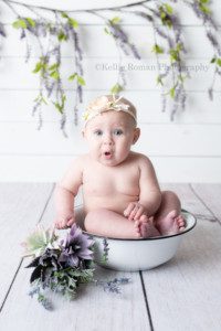 happiest six month old a little girl sitting in a white vintage tub in a milwaukee Wisconsin photo studio. The baby is a surpised expression on her face and is wearing only a diaper that you can't see. She has an ivory headband on and there are purple and green succulents next to the white tub. The baby is in front of a white wood wall with a lilac vine behind her.
